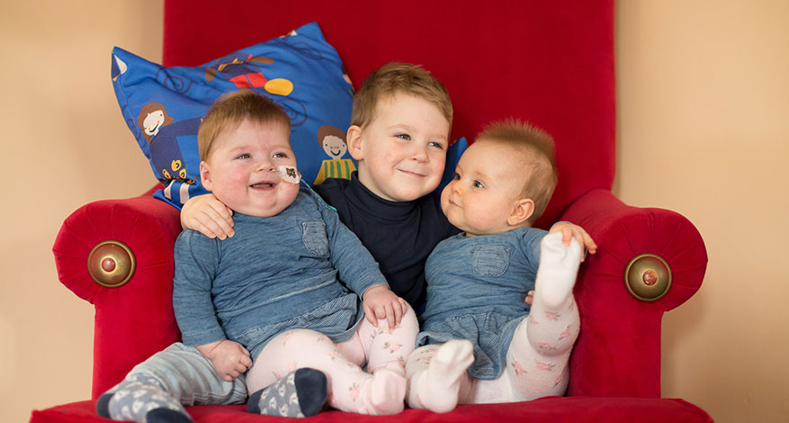 3 children sit on large red chair at Demelza